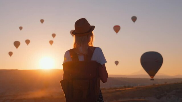 beautiful woman in hat backpack walking towards fairytale view balloons at sunrise in Gorem Cappadocia Turkey. Image of traveler and wanderer, she looks at fabulous and beautiful sunrise in mountains