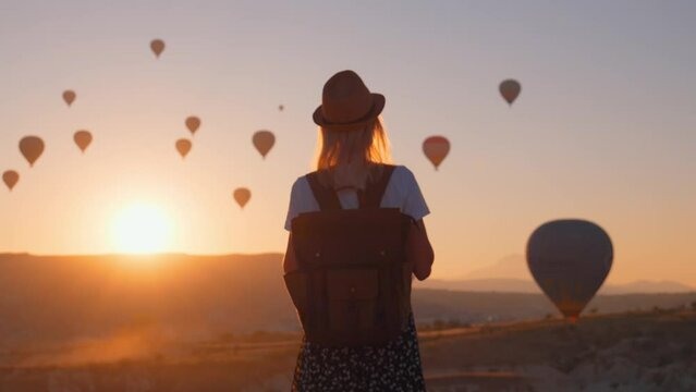 Young beautiful blonde woman traveler with backpack and hat walking and enjoying hot air balloon rides on Goreme hill, Cappadocia, Turkey at dawn in sunshine of girl. Hiking in fairy tale