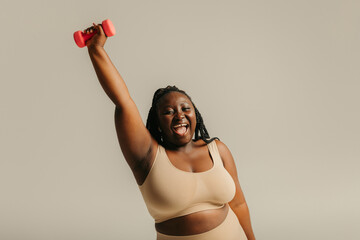 Joyful plus size African woman in sportswear exercising with dumbbell on studio background