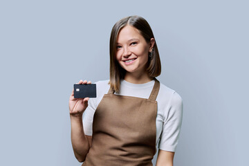Young happy woman service worker in apron with credit card in hand, on gray background