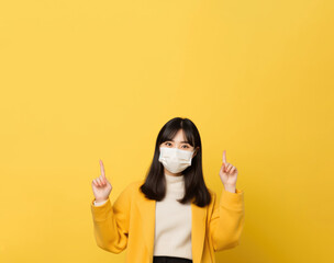 young asian woman in medical mask points finger up on yellow background. coronavirus protection and prevention concept
