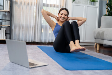Fototapeta na wymiar Asian woman in sportswear doing crunch on exercising mat as home workout training routine. Attractive girl engage in her pursuit of healthy lifestyle with online exercise training video. Vigorous