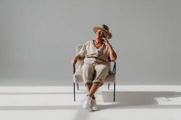 Relaxed gray hair senior woman in elegant hat sitting in comfortable chair at the sunny studio