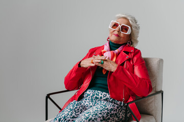 Fashionable senior woman in red leather jacket sitting in comfortable chair on grey background