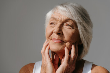 Portrait of thoughtful senior woman touching face and smiling against grey background