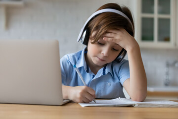 Focused 8s boy in headphones studying online or making homework at home, serious child kid taking notes, listening to lecture or watching webinar, homeschooling and distance learning concept