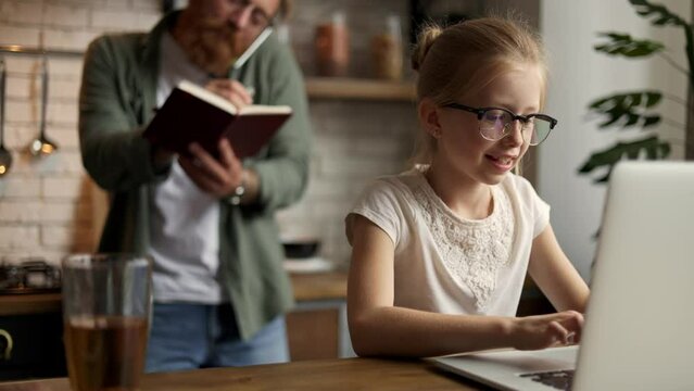 Child using laptop near blurred busy father talking on smartphone in kitchen, slow motion