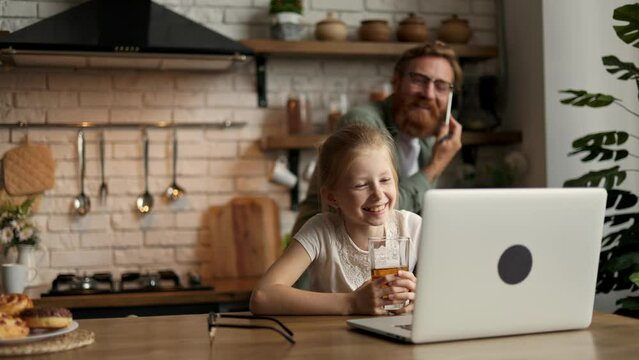 Smiling child with juice using laptop while father talking on smartphone in kitchen at home