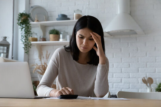 Unhappy stressed Asian woman checking bills, calculating taxes or household expenses, sitting at kitchen table with laptop, frustrated young female tenant renter having money problem, planning budget
