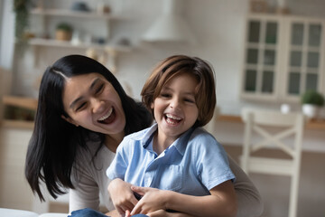 Overjoyed Asian mother and little son hugging and laughing, spending leisure time at home together,...
