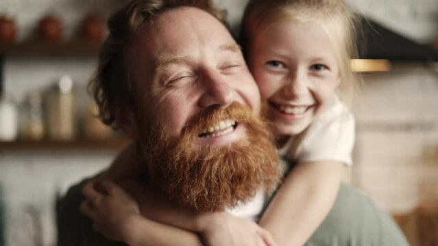 Portrait of girl hugging cheerful bearded father and looking at camera at home, slow motion