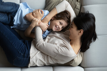 Top view smiling loving Asian mother and son hugging, family relaxing lying on couch at home, happy caring young mom with 7s boy child enjoying tender moment, having fun, motherhood concept