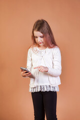 Pensive kid girl in white using texting sms at mobile phone posing at beige backdrop, think look. Child model 7 year old with cellphone. Children communication, education concept. Copy ad text space