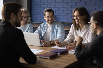 Five happy employees sit at table in boardroom, having pleasant informal conversation, discuss project at meeting or brainstorming. Colleagues enjoy good work relations, have fun, joking, negotiating