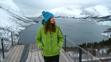 Lofoten islands, Senja region, Norway. Winter. Young girl in green jacket and blue hat stands on...