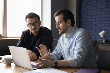 Two young focused business partners sit at desk in modern office discuss project details, share ideas, mentor teaches employee new business application, explains online work. Collaboration, teamwork