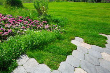 Park landscape design with flowerbeds and green lawn by the granite tile sidewalk, spring sunny greenery landscape.