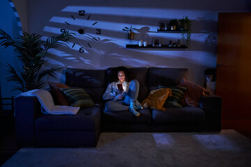 Naklejka premium Young woman with serious expression looking at cell phone sitting in living room at night. Concentrated latina female using mobile on sofa at home. Copy space, generation z and free time indoors.