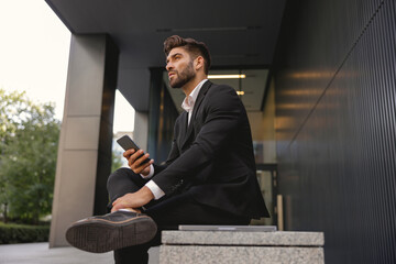 Businessman with mobile phone sitting on bench on office building background. High quality photo