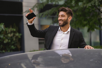 Smiling male manager standing near car with phone and waving hi to colleagues. High quality photo