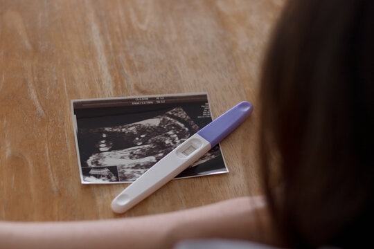 Happy pregnant woman looking at ultrasound pictures with pregnancy test on a wooden background. Result of ultrasonography for baby and mother healthcare checkup. Gynecology and Pregnancy care concept.