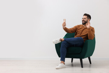 Handsome man having video chat via smartphone while sitting in armchair near white wall indoors, space for text