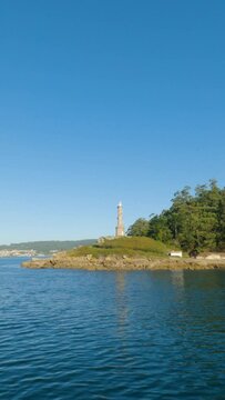 Sailing past Tenlo Chico Peninsula, Tambo Island. Lush trees meet rocky shores under a calm sea. Combarro in the distance. Tenlo Chico lighthouse and blue summer sky reflected on the calm sea.