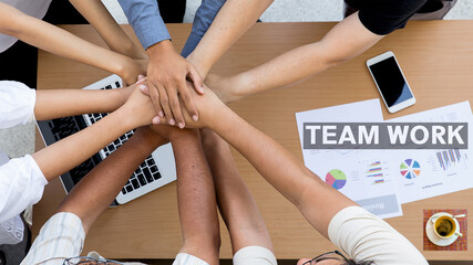 Grupo de trabajo uniendo sus manos mostrando la importancia de trabajar en equipo. Se vizualiza el grupo unido, su mesa de trabajo, la laptop, taza de café, papeles de trabajo. Texto Team Work.