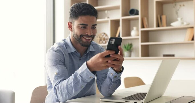 Young 30s smiling Moroccan office employee hold cellphone, sit at workplace, typing message use application, enjoy on-line personal communication, take break, have fun with new e-dating app services