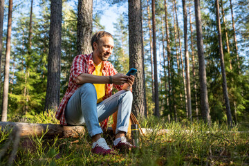 Happy middle aged man browsing in smartphone sitting on log in park forest. Guy enjoying nature looking at phone screen resting outdoors, relaxing watching social media, funny videos, surfing online.