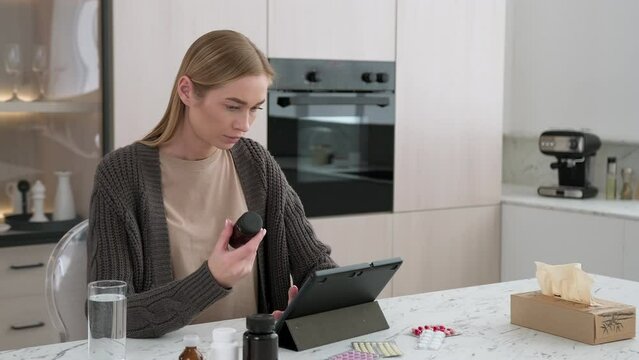 A woman reads instructions for using pills using a gadget while sitting at a table in the kitchen.