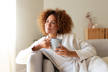 Pensive multiracial woman relaxing at home, sitting on the sofa drinking tea.
