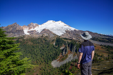 Adventurous athletic male hiker standing on top of a hill looking at Mount Baker on a beautiful sunny fall day in the Pacific Northwest.
