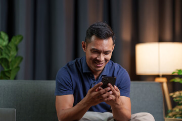 A strong Asian dark-skinned man sits on a sofa holding a smartphone, checking emails online,...