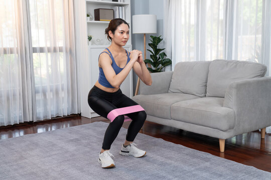 Vigorous energetic woman doing exercise at home with resistance sport band for leg muscle gain. Young athletic asian woman strength and endurance training session as home workout routine with squat.