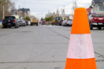 a slightly worn orange traffic cone placed to close a street where they work with machines and trucks in the background in blur