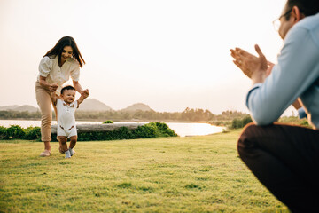 A happy mother and father helping their toddler take her first steps in the green grass of the park. baby learning concept