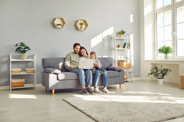 Full length photo of a happy young family with little child girl sitting on the sofa using modern laptop together. Parents resting on couch watching funny cartoon online or talking on video call.