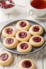 Thumbprint a buttery and crumbly vanilla cookie dough filled with delicious and tart raspberry jam closeup on the plate served with tea on the white wooden table. Vertical