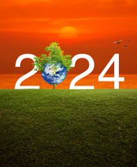 2024 white text with planet and tree on green grass field over sunset sky and sea, Happy new year...