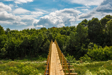 Fototapeta na wymiar Colorful rural landscape with a view of a wooden footbridge over a river against the backdrop of clouds floating across the sky