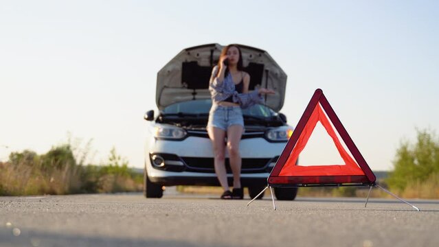 Road side warning triangle, warning oncoming traffic of a broken down car, with a woman using her cell phone to call for assistance