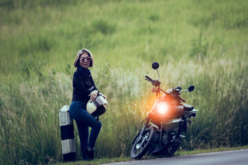 pretty woman holding safety helmet standing beside small enduro motorcycle against green grass field