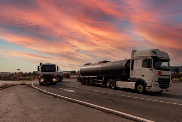 Two tankers with dangerous goods, one parked and one driving under a dramatic sky at dawn.