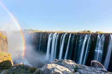 Panorama photo of Victoria Falls waterfall on Zambezi river in very high flow in late evening light with rising spray and intensive rainbow over falls.