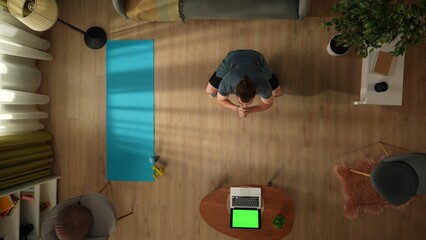 Top view capturing a young man doing squats in front of the laptop with an advertising area, workspace mock up on it, as if he is watching a video. Home, room, indoor activities.