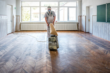 Sanding hardwood floor with the grinding machine. Repair in the apartment. Carpenter doing parquet wood floor polishing maintenance work by grinding machine - obrazy, fototapety, plakaty