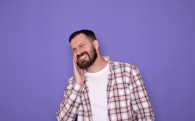 Dental problems. Portrait of unhealthy man pressing sore cheek, suffering acute toothache, periodontal disease, cavities or jaw pain. Indoor studio shot isolated on purple background. 