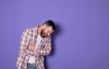 Young bearded man having a stomachache. Young man keeping hands at his stomach because painful disease feeling unwell on purple background. Ache concept.