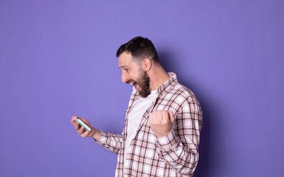 Portrait of extremely happy handsome bearded man gamer playing video game on mobile phone, celebrating victory, complete level on purple background. - Powered by Adobe